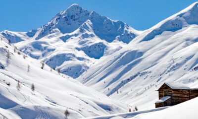 Baita di legno in primo piano immersa nella neve con delle montagne completamente innevate sullo sfondo con un cielo privo di nuvole