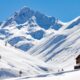 Baita di legno in primo piano immersa nella neve con delle montagne completamente innevate sullo sfondo con un cielo privo di nuvole