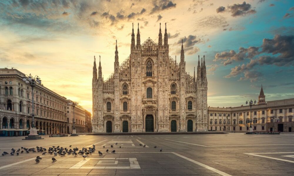 Sagrato di piazza del Duomo di Milano all'alba senza persone con dei piccioni in primo piano e altri edifici storici ai lati della piazza