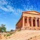Tempio greco nella Valle dei Templi ad Agrigento con erba bruciata dal sole alla base, una pianta al lato del tempio in una giornata di sole