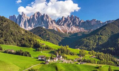 Piccolo agglomerato di case con una chiesetta nei prati verdi di montagna con abeti con sullo sfondo delle montagne rocciose con delle nuvole bianche nel cielo