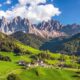 Piccolo agglomerato di case con una chiesetta nei prati verdi di montagna con abeti con sullo sfondo delle montagne rocciose con delle nuvole bianche nel cielo