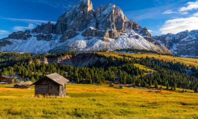 Valle di montagna con delle baite sparse in primo piano nei prati, degli abeti nelle vicinanze e sullo sfondo delle montagne rocciose con della neve sul costone