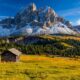 Valle di montagna con delle baite sparse in primo piano nei prati, degli abeti nelle vicinanze e sullo sfondo delle montagne rocciose con della neve sul costone