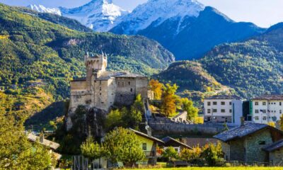 Prato verde di montagna con case, alberi e un castello medievale in Valle d'Aosta con la sua torre e le guglie con sullo sfondo delle montagne con alberi e delle altre cime innevate dietro