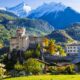 Prato verde di montagna con case, alberi e un castello medievale in Valle d'Aosta con la sua torre e le guglie con sullo sfondo delle montagne con alberi e delle altre cime innevate dietro