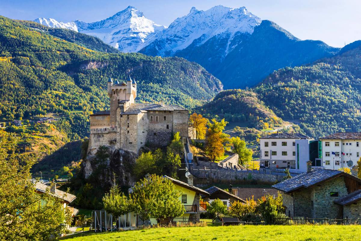 Prato verde di montagna con case, alberi e un castello medievale in Valle d'Aosta con la sua torre e le guglie con sullo sfondo delle montagne con alberi e delle altre cime innevate dietro