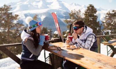 Una ragazza e un ragazzo con equipaggiamento da sci che si guardano seduti ad un tavolo di legno mentre bevono e mangiano con sullo sfondo degli abeti e delle montagne con le cime piene di neve