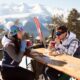 Una ragazza e un ragazzo con equipaggiamento da sci che si guardano seduti ad un tavolo di legno mentre bevono e mangiano con sullo sfondo degli abeti e delle montagne con le cime piene di neve