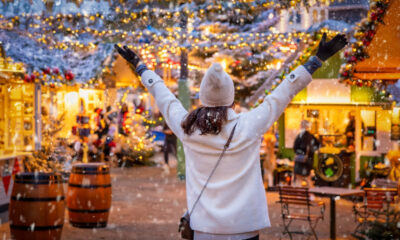 Donna felice vestita di bianco con un berretto di lana in testa, vista di spalle, tra le casette di un mercatino di Natale