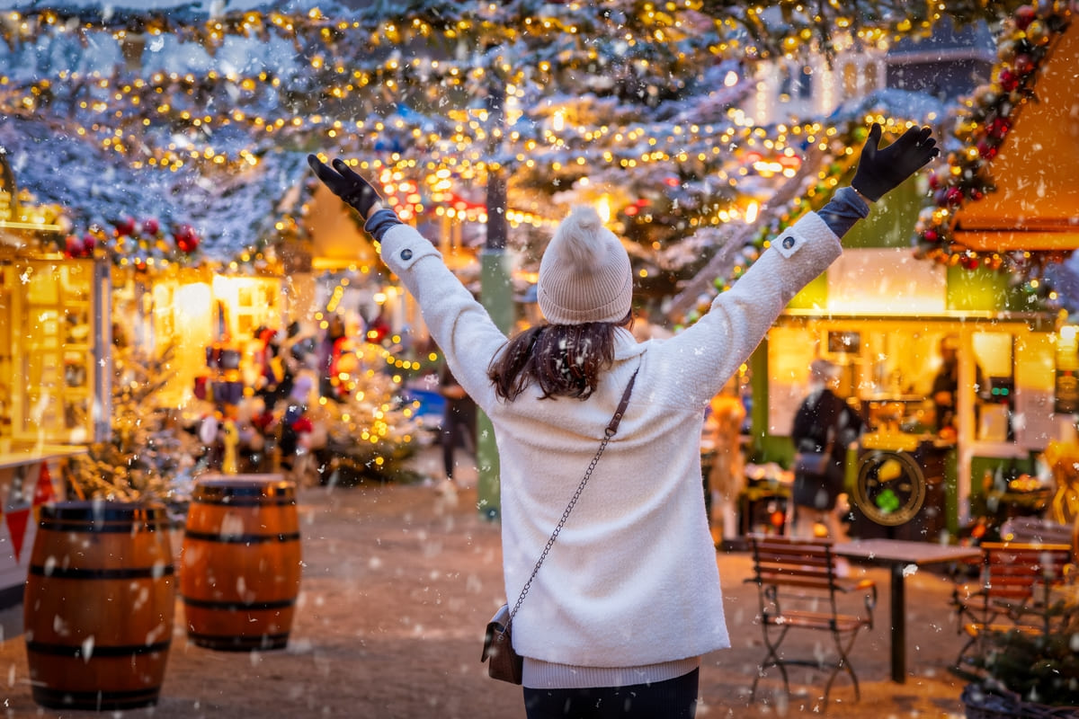 Donna felice vestita di bianco con un berretto di lana in testa, vista di spalle, tra le casette di un mercatino di Natale