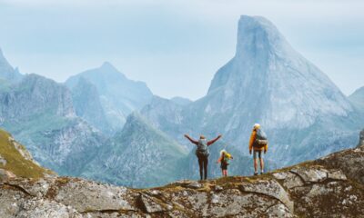 Mamma, papà e figlia di spalle equipaggiati per fare trekking con zaino in spalla sul costone roccioso di una montagna mentre guardano la vallata con delle montagne sullo sfondo