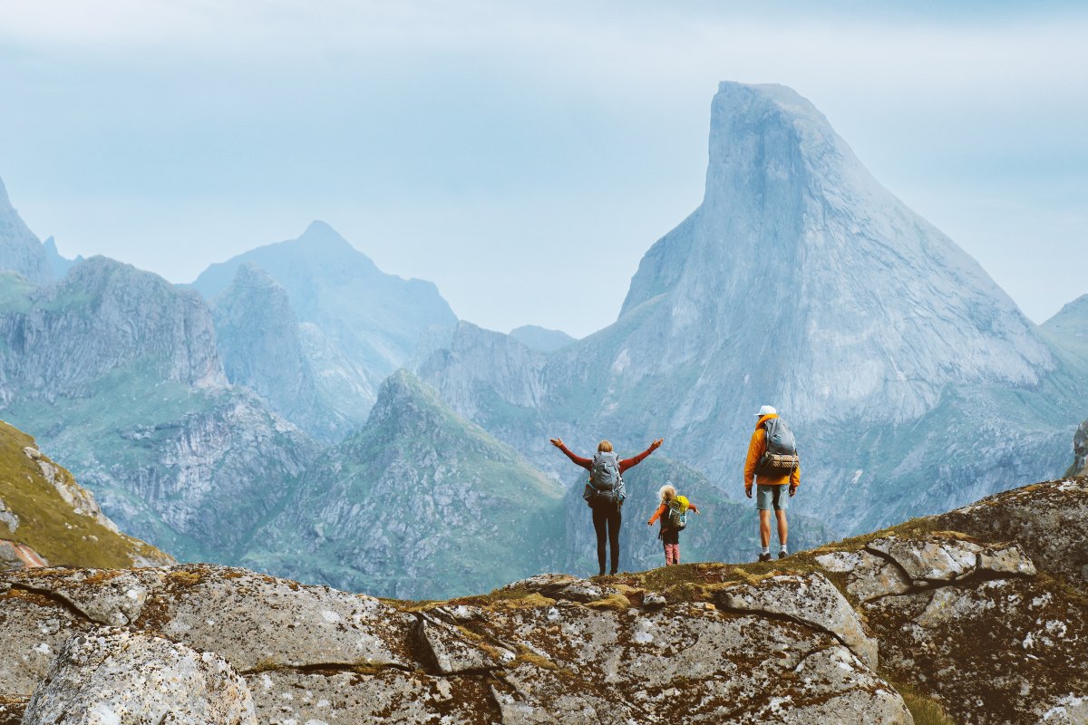 Mamma, papà e figlia di spalle equipaggiati per fare trekking con zaino in spalla sul costone roccioso di una montagna mentre guardano la vallata con delle montagne sullo sfondo