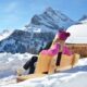 Ragazza seduta con cappello e giacca rossa seduta su una panchina di legno che prende il sole in montagna con tanta neve in primo piano e sullo sfondo delle cime di montagne innevate