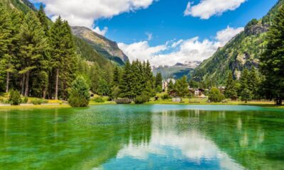 Specchio d'acqua con sfumature verdi in una valle alpina con degli abeti sulle sponde, un piccolo ponte di legno con sullo sfondo delle case e altre montagne immerse nelle nuvole bianche
