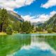 Specchio d'acqua con sfumature verdi in una valle alpina con degli abeti sulle sponde, un piccolo ponte di legno con sullo sfondo delle case e altre montagne immerse nelle nuvole bianche