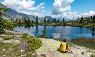Ragazzo con lo zaino giallo in spalla seduto su una roccia sulla riva di un lago alpino circondato da abeti che guarda il panorama