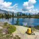 Ragazzo con lo zaino giallo in spalla seduto su una roccia sulla riva di un lago alpino circondato da abeti che guarda il panorama