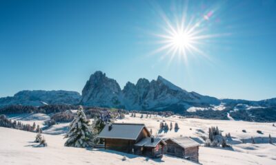 Baita di legno al centro di una vallata innevata con abeti e sullo sfondo delle cime rocciose anch'esse innevate con il sole alto nel cielo