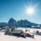 Baita di legno al centro di una vallata innevata con abeti e sullo sfondo delle cime rocciose anch'esse innevate con il sole alto nel cielo