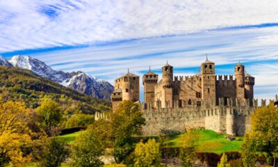 Mura e torri di un castello medievale circondato da prati e alberi con sullo sfondo delle montagne con le cime innevate in una giornata di autunno al tramonto