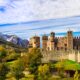 Mura e torri di un castello medievale circondato da prati e alberi con sullo sfondo delle montagne con le cime innevate in una giornata di autunno al tramonto