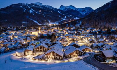 Case e chalet di un borgo di montagna illuminati dalle luci artificiali di sera immersi nella neve con sullo sfondo delle montagne con abeti e neve