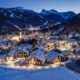 Case e chalet di un borgo di montagna illuminati dalle luci artificiali di sera immersi nella neve con sullo sfondo delle montagne con abeti e neve