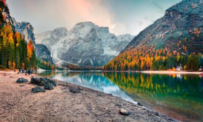 Piccola spiaggia di sassi che da su un lago di montagna con degli escursionisti che camminano lungo la riva con ai lati delle montagne con arbusti dalla foglie arancio e verdi e sullo sfondo altre montagne innevate