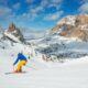 Sciatore con giacca blu e pantaloni gialli che affronta una pista da sci con delle montagne sullo sfondo