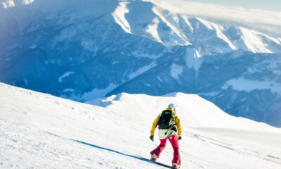 Ragazzo con casco bianco, zaino, giacca gialle e pantaloni rossi che fa snowboard su una pista da sci con sullo sfondo delle montagne innevate in una giornata di sole