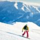 Ragazzo con casco bianco, zaino, giacca gialle e pantaloni rossi che fa snowboard su una pista da sci con sullo sfondo delle montagne innevate in una giornata di sole