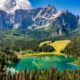 Abeti che circondano un lago con un prato verde e delle montagne delle Alpi sullo sfondo in una giornata di sole
