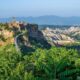 Città vecchia di Civita di Bagnoreggio in cima alla montagna con il suo ponte che porta al borgo, della vegetazione in primo piano e sullo sfondo altri monti del Lazio