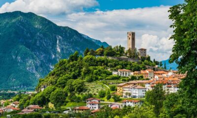 Case alla base di una collinetta con della vegetazione in primo piano e sulle pareti del colle con una torre antica in cima e sullo sfondo delle montagne