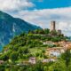 Case alla base di una collinetta con della vegetazione in primo piano e sulle pareti del colle con una torre antica in cima e sullo sfondo delle montagne