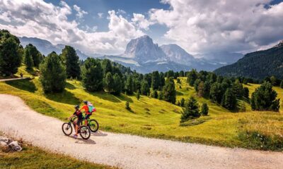 Coppia in gita in bicicletta in montagna su un sentiero sterrato con prati verdi e abeti con sullo sfondo delle montagne rocciose