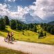 Coppia in gita in bicicletta in montagna su un sentiero sterrato con prati verdi e abeti con sullo sfondo delle montagne rocciose